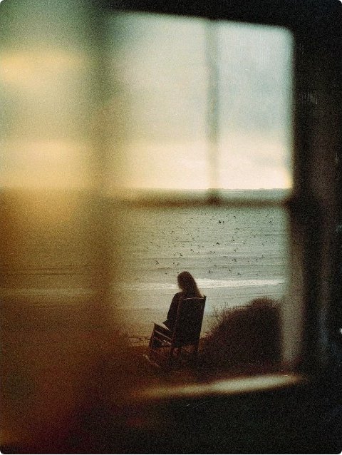 A lone figure in a chair looking out at a hazy shoreline through a window
