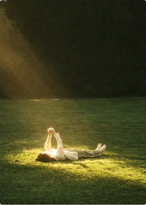 A person lying in a sunlit patch of grass, arms raised toward the light