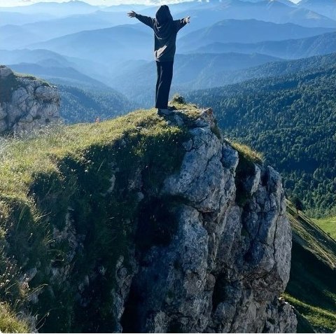 A person standing arms-wide on a mountain summit above blue ridgelines
