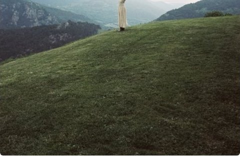 A figure standing arms-open on a green hillside beneath misty mountains